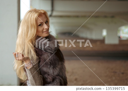 Woman stands under structure with long hair and fur piece on shoulders, casual pose and expression Woman stands under structure with long hair and fur piece on shoulders, casual pose and expression 135399715