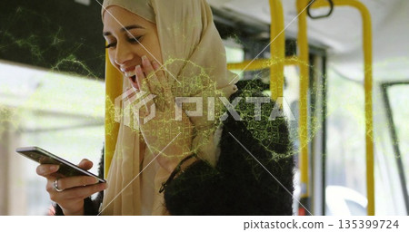 Smiling woman wearing hijab and black jacket, holding phone inside bus by windows with yellow poles 135399724