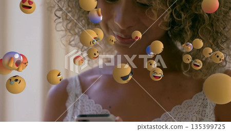 Holding smartphone, woman wearing lace top gazing at device at home, with floating emoji spheres 135399725