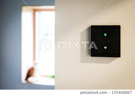 Modern smart home wall control panel with illuminated touch icons on a white wall. Minimalist interior design featuring a black electronic switch for lighting and home automation.  135400067