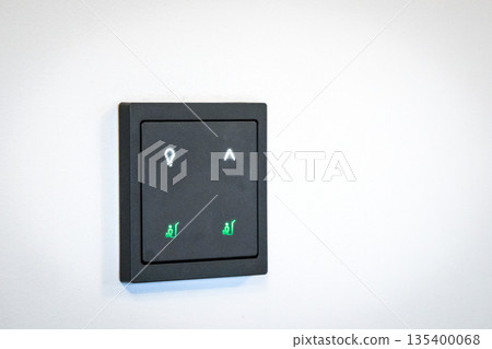 Modern smart home wall control panel with illuminated touch icons on a white wall. Minimalist interior design featuring a black electronic switch for lighting and home automation. Modern smart home wall control panel with illuminated touch icons on a white wall. Minimalist interior design featuring a black electronic switch for lighting and home automation. 135400068