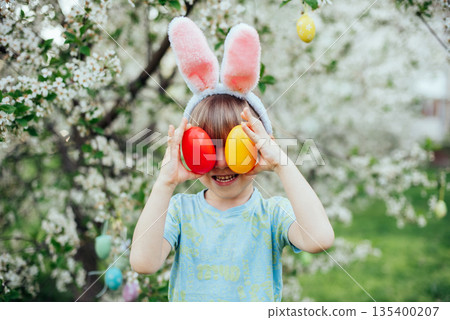 Easter Egg Hunt with Child Carrying Bright Plastic Eggs. Smiling child wearing bunny ears in a blooming spring garden Easter Egg Hunt with Child Carrying Bright Plastic Eggs. Smiling child wearing bunny ears in a blooming spring garden 135400207