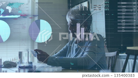 Senior woman holding smartphone and computer mouse in home office, reviewing data graphics 135400383