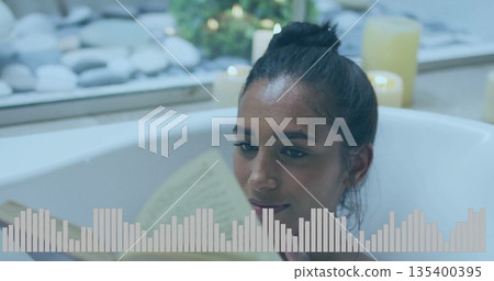 Relaxing South Asian woman reading paperback book in bathtub by window, with stones and lit candles 135400395