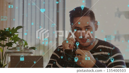 Asian man holding smartphone and scrolling icons on laptop in home office, with plant and jar 135400503