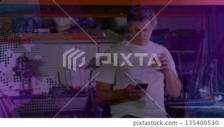 Leaning mature technician holding smartphone and coffee in workshop, shelving and digital overlays Leaning mature technician holding smartphone and coffee in workshop, shelving and digital overlays 135400530
