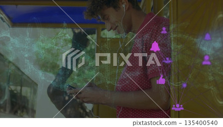 Leaning man wearing wired earphones checking smartphone at yellow bus shelter, with overlay icons 135400540