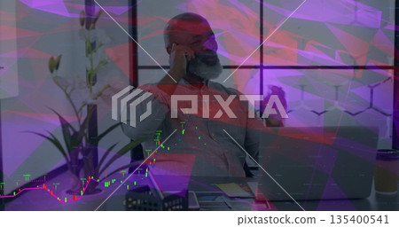 Mature man talking on smartphone and gesturing at modern office desk with laptop, coffee cup, plant 135400541