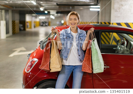 A smiling woman stands next to her car in a parking garage, holding several shopping bags A smiling woman stands next to her car in a parking garage, holding several shopping bags 135401519