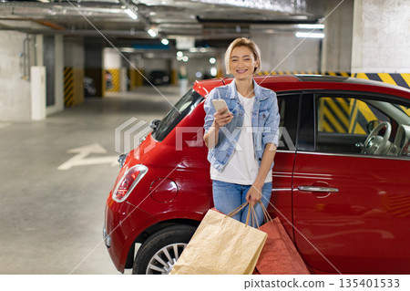 A smiling woman stands next to her red car in a parking garage, holding shopping bags and a phone 135401533