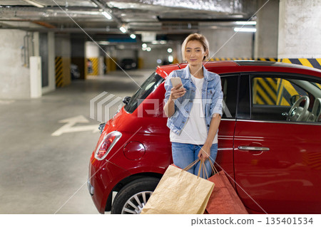 A woman stands in a parking garage with shopping bags and a smartphone next to her red car A woman stands in a parking garage with shopping bags and a smartphone next to her red car 135401534