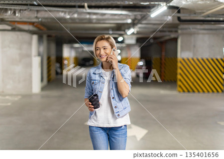 A smiling woman walks through a parking garage while talking on her phone and holding a coffee cup A smiling woman walks through a parking garage while talking on her phone and holding a coffee cup 135401656