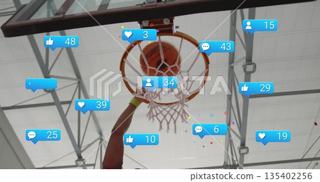 Slam dunking basketball player driving ball through court hoop, with floating social media icons 135402256