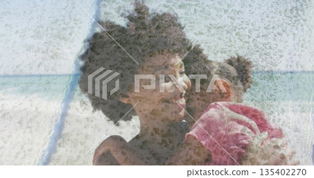 Lifting mother hugging daughter at sandy beach shoreline, displaying curly hair and pink T-shirt 135402270