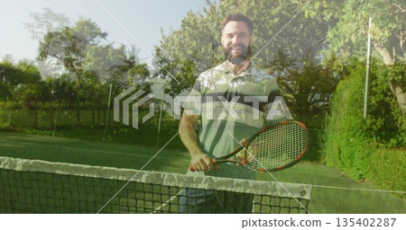 Smiling male tennis player holding racket behind net on grass court, with net post and hedges 135402287