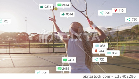 Serving woman wearing white top gripping racket tossing ball on tennis court, digital overlay icons 135402296