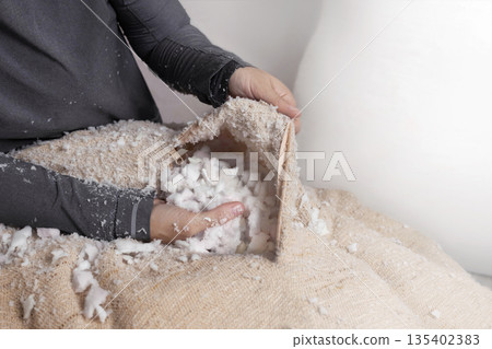 Woman fills a pillow case with foam rubber. Foam pieces, crumbs. Woman repairs furniture pillows. White polyurethane foam rubber pieces. stuffing for pillows and furniture. Cropped photo 135402383