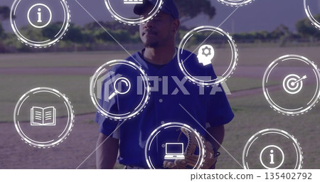 Standing baseball player holding leather glove wearing blue cap on dirt infield, with digital icons 135402792