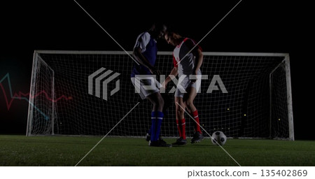 Challenging two opposing soccer players contesting ball on grass pitch under floodlights, goal net Challenging two opposing soccer players contesting ball on grass pitch under floodlights, goal net 135402869