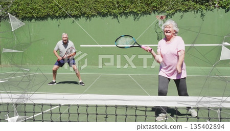 Swinging senior woman in pink shirt returning shot on tennis court, rackets, net, hedge, copy space 135402894