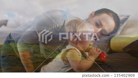Leaning mother watching daughter mouthing red green teething toy in bedroom, with neutral sheets Leaning mother watching daughter mouthing red green teething toy in bedroom, with neutral sheets 135402909