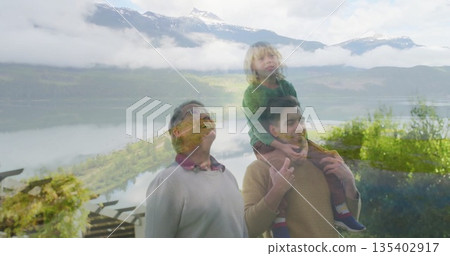 Standing family trio facing mountain vista over lake on deck, with wooden railing and pergola beams 135402917