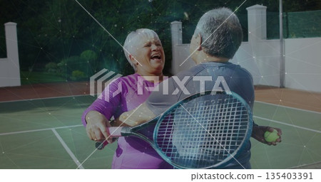 Laughing senior couple in sportswear hugging on tennis court, holding rackets and ball 135403391