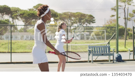 Two training partners in white attire holding rackets on tennis court with bench, bag, fence Two training partners in white attire holding rackets on tennis court with bench, bag, fence 135403470