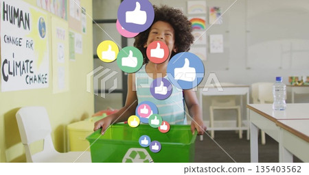 Holding rim of green recycling bin, boy standing in school classroom, with digital thumbs-up icons 135403562