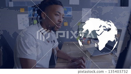 Man wearing white T-shirt and headphones typing on keyboard at desk with map overlay, copy space 135403732