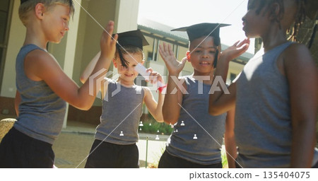 Celebrating four children wearing uniforms in school courtyard with mortarboard hats and diploma Celebrating four children wearing uniforms in school courtyard with mortarboard hats and diploma 135404075