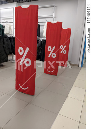 Three red signs with a smiling face and percentage symbol are placed in a store aisle next to clothing racks during a sales event in the afternoon Three red signs with a smiling face and percentage symbol are placed in a store aisle next to clothing racks during a sales event in the afternoon 135404124