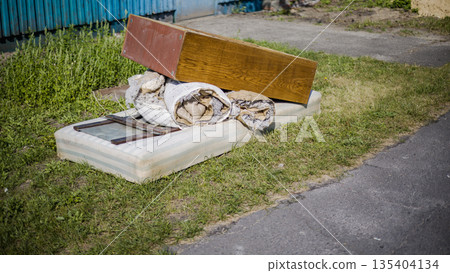 Several pieces of old furniture and materials are piled on a roadside next to a blue fence in a grassy area during daytime hours 135404134