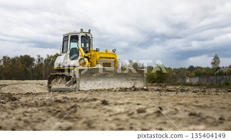 A large construction vehicle pushes soil across a construction site under a cloudy sky. Trees are visible in the background, marking the area 135404149