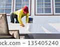 A painter applying fresh white paint to an exterior wall with a roller. 135405240