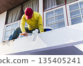 Worker painting an exterior wall with a roller during house maintenance. 135405241