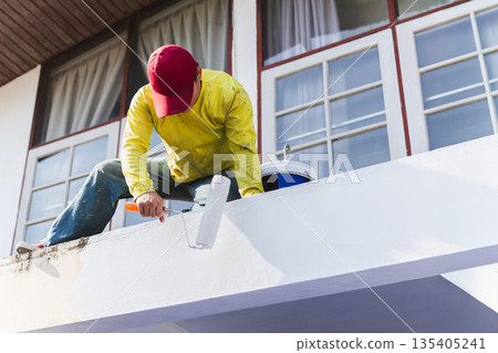 Worker painting an exterior wall with a roller during house maintenance. Worker painting an exterior wall with a roller during house maintenance. 135405241