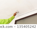 A worker applying plaster with a putty knife to smooth a ceiling surface. 135405242