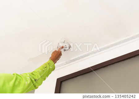 A worker applying plaster with a putty knife to smooth a ceiling surface. 135405242