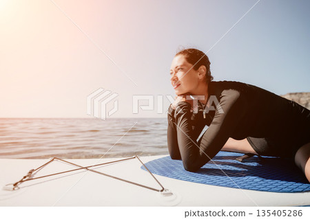 Woman Paddleboard Ocean Relaxing - A woman wearing a wetsuit relaxes on a paddleboard on the ocean while looking out at the horizon. 135405286