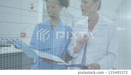 Two medical staff standing in hospital hallway, reviewing patient chart with digital trend overlay 135405372