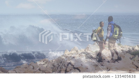 Holding hands couple standing on rocky coastal outcrop, with hiking backpacks and boots, copy space 135405638