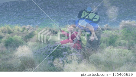 Kneeling woman in red jacket pointing and man hiking hillside, carrying green backpack and poles 135405639
