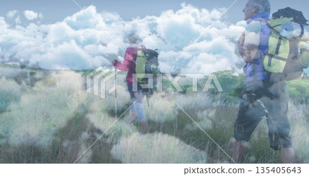 Traversing hikers wearing jackets across field under cloudy sky, carrying poles and packs with pads Traversing hikers wearing jackets across field under cloudy sky, carrying poles and packs with pads 135405643