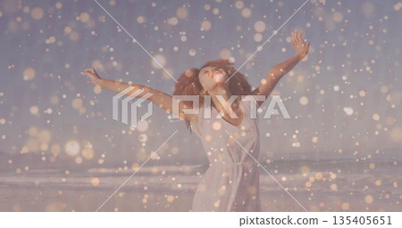 Spreading arms woman in white summer dress greeting ocean waves at sandy beach, with golden bokeh 135405651
