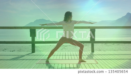 Performing yoga pose adult woman in patterned leggings on seaside deck, with coral mat and railing 135405654