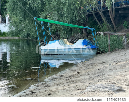 Pedal boat moored by a lake with green trees on a sandy shore. Pedal boat moored by a lake with green trees on a sandy shore. 135405683