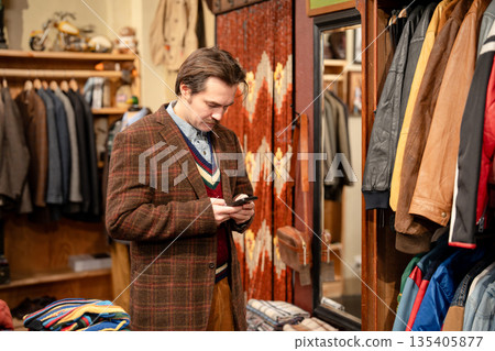 Elegant man in classy blazer checking smartphone during shopping in vintage men's clothing store. 135405877