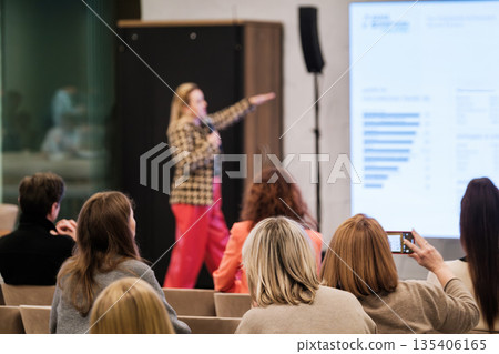 Speaker presents at conference with audience listening and recording during presentation in a modern venue Speaker presents at conference with audience listening and recording during presentation in a modern venue 135406165