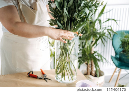 Professional florist places tall green stems into glass vase at workbench. Floral design process, creative workspace, artisanal skills, flower workshop. Professional florist places tall green stems into glass vase at workbench. Floral design process, creative workspace, artisanal skills, flower workshop. 135406170